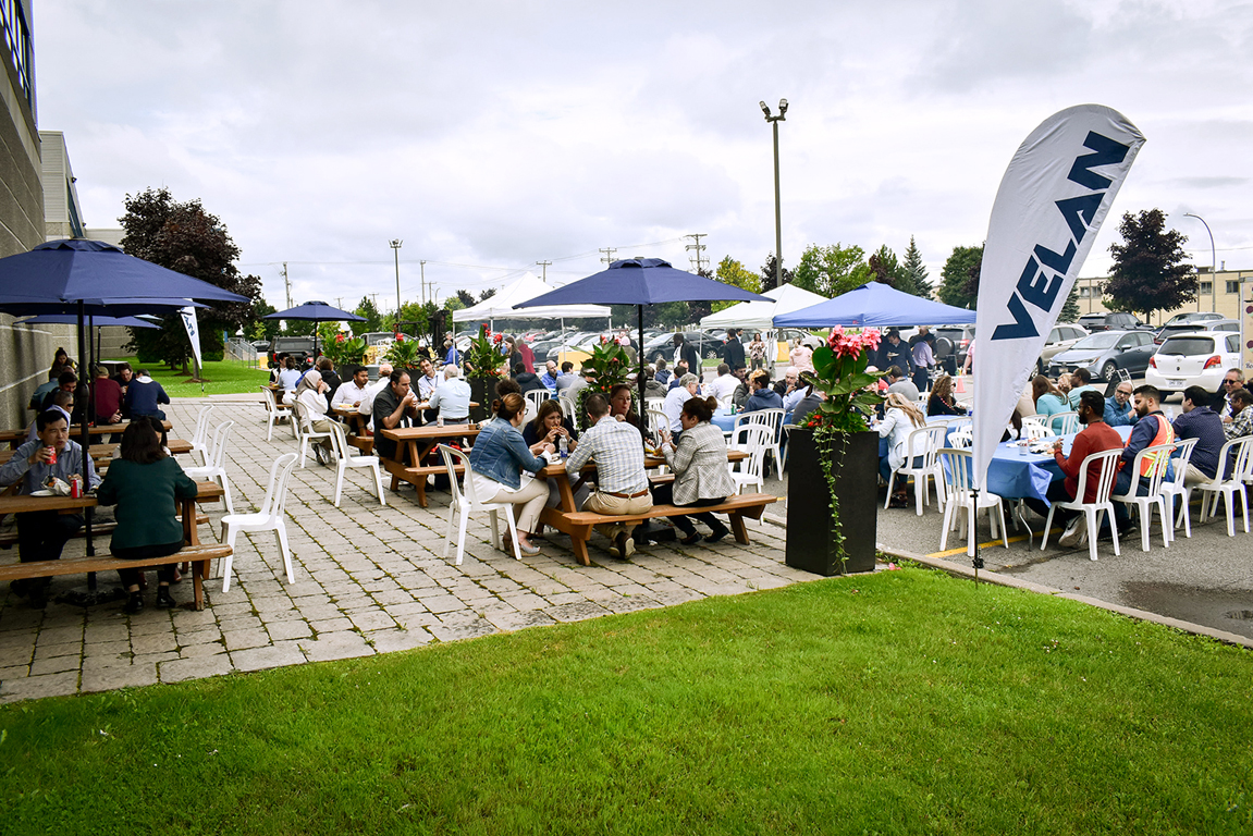 A lively outdoor gathering with people seated at picnic tables under blue umbrellas on a brick patio. A white flag with the word "VELAN" stands prominently near the seats. The background includes green grass, trees, and parked cars. The sky is overcast.