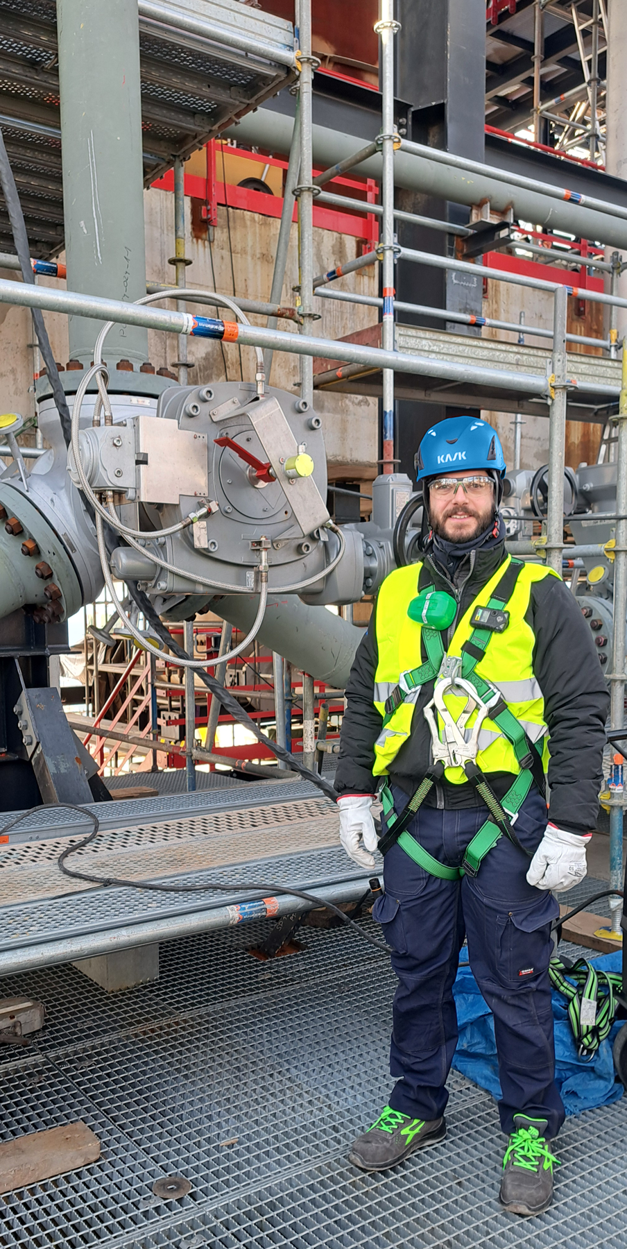 A construction worker wearing safety gear, including a blue helmet, high-visibility vest, gloves, and a harness, stands on a metal platform. Behind them is industrial scaffolding and machinery.