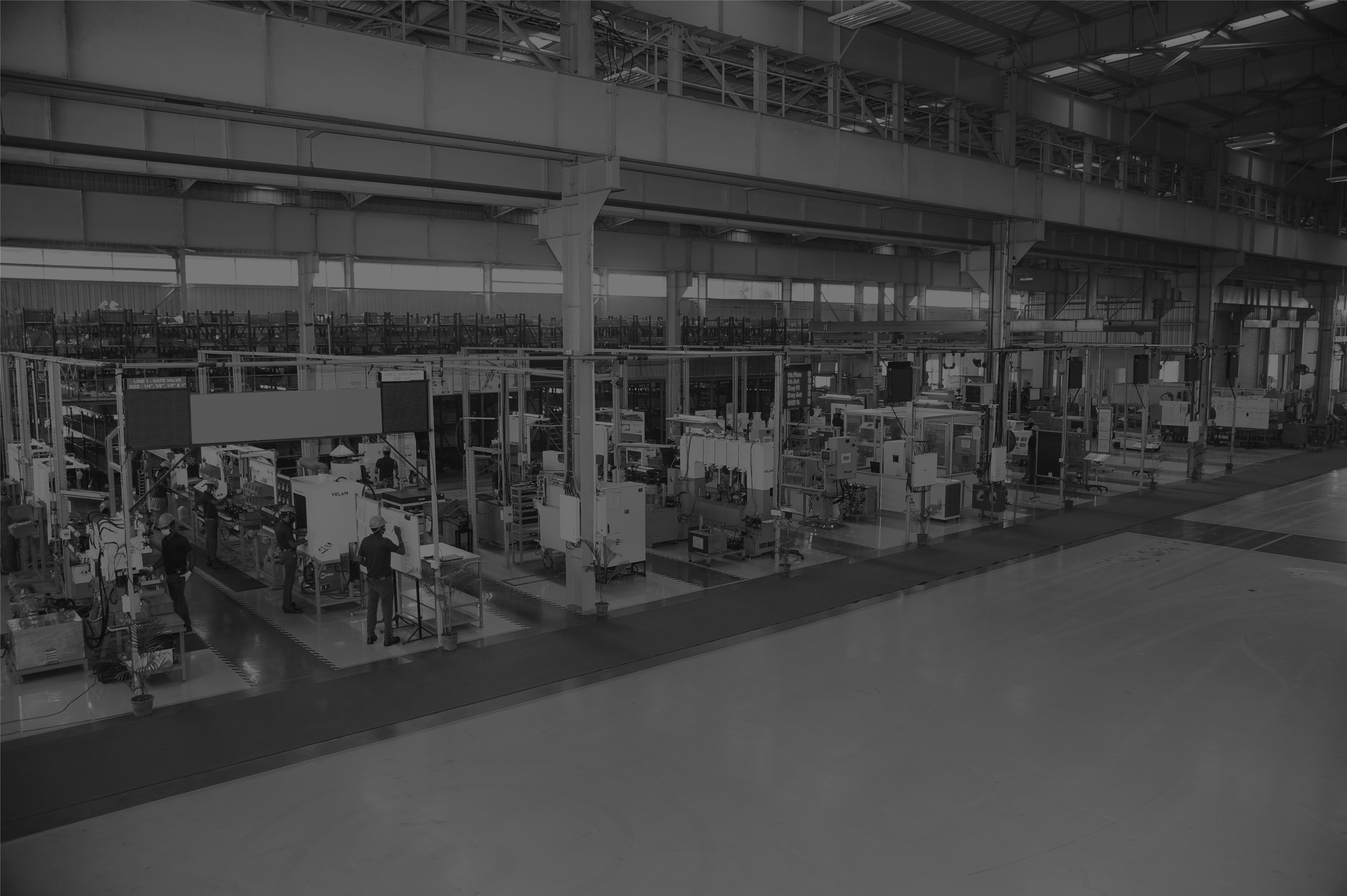 Black and white image of an expansive, modern industrial manufacturing facility. Various machines and equipment are organized in rows, with a few workers attending to the machinery. The space is well-lit, with high ceilings and structural beams visible.