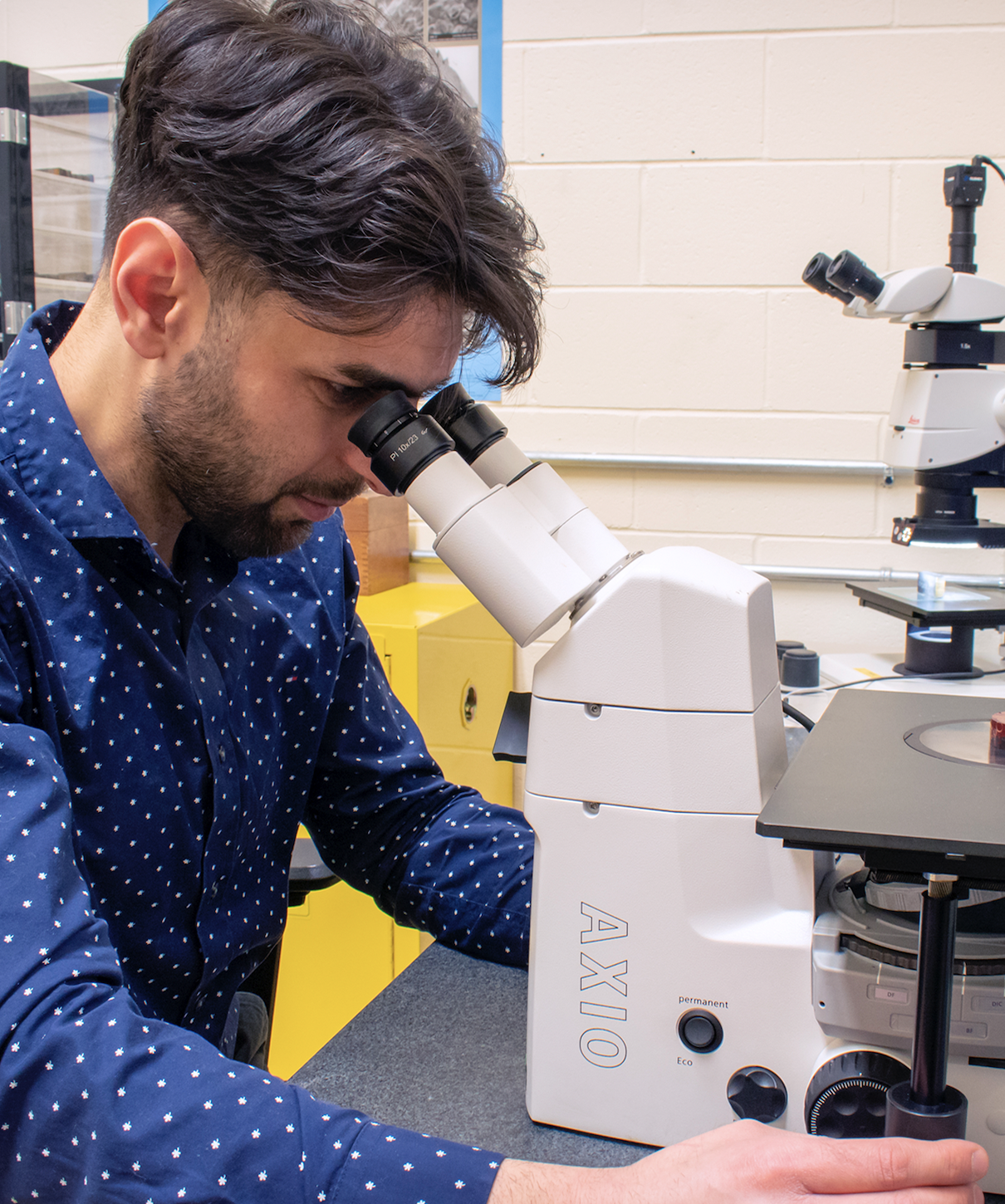 A man in a navy polka dot shirt is looking through a microscope labeled "AXIO" in a laboratory setting. The background features another microscope and laboratory equipment on a counter.