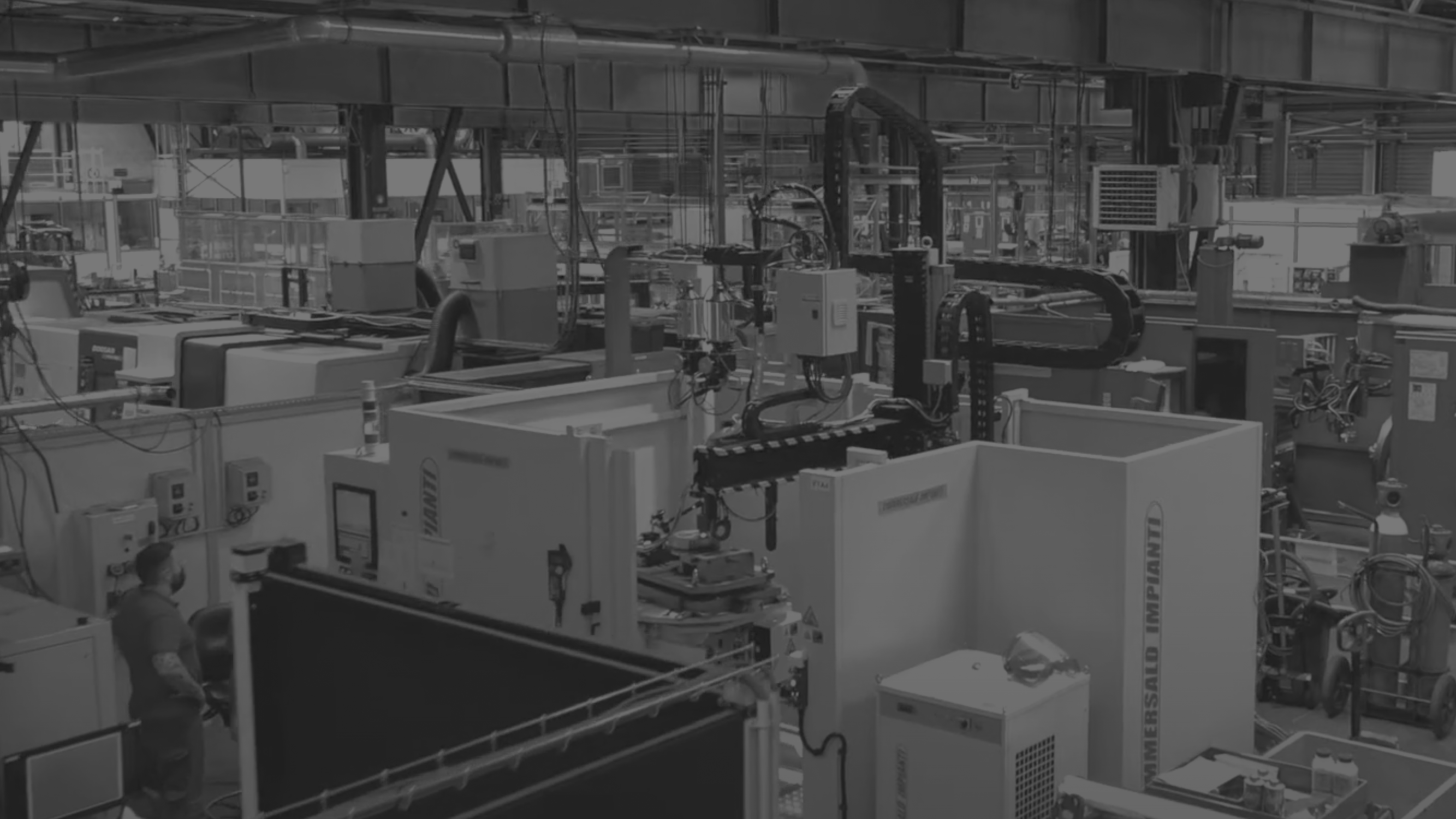 A black and white image of an industrial manufacturing facility. Various machines, equipment, and control panels are arranged throughout the space. A worker wearing a helmet is standing on the left side, operating one of the machines.