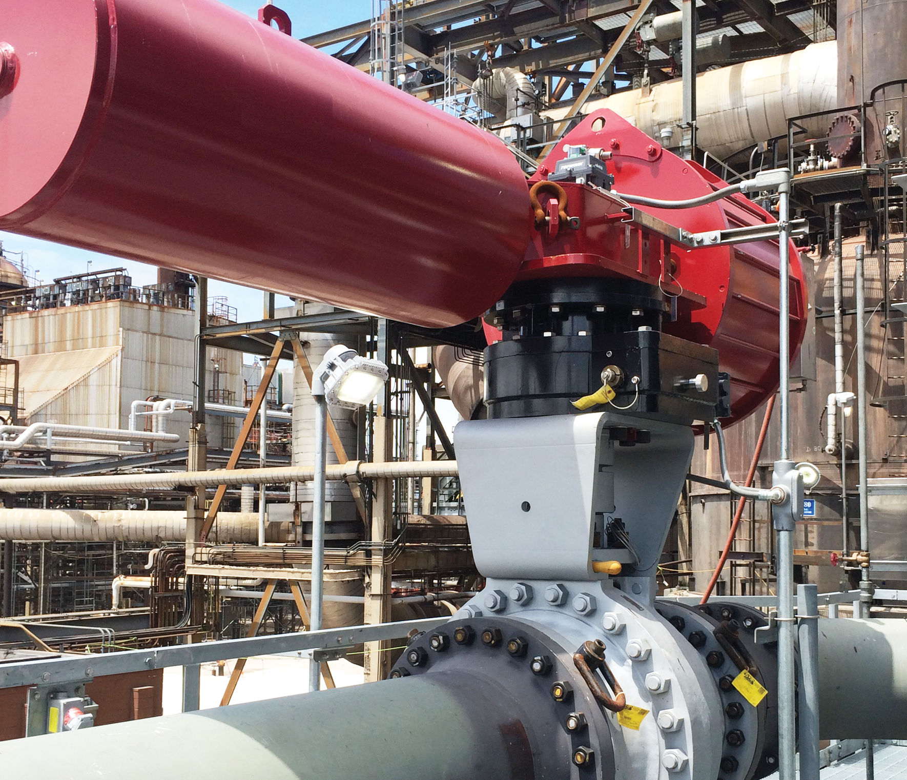 An industrial setting featuring a large valve actuator connected to a pipeline. The actuator is red and sits on a gray valve with numerous bolts. Background reveals complex network of pipes and industrial structures.