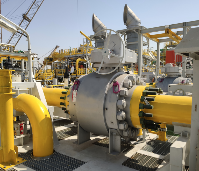 A large industrial pipeline system with a prominent spherical valve and yellow pipes, surrounded by complex metal structures and machinery at an outdoor facility under a clear sky.